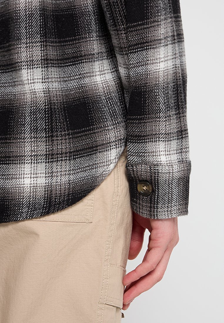 Schwarz-weiße kariertes Hemd mit Knopfleiste, abgerundetem Saum, langen Ärmeln und einem einzelnen Knopf an den Handgelenken.