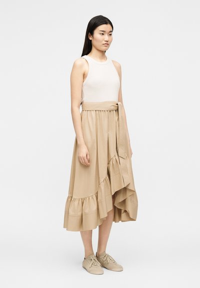 Νεαρή γυναίκα που φοράει λευκό τοπ χωρίς μανίκια συνδυασμένο με μπεζ μίντι φούστα με βολάν και ψηλή μέση και μπεζ αθλητικά, στέκεται μπροστά σε απλό φόντο.