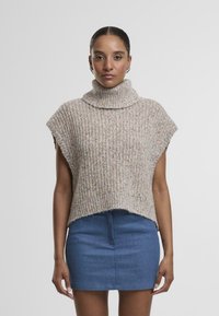 Poncho en tricot côtelé beige, col roulé, manches courtes, porté sur une mini-jupe en denim bleu. L'ourlet est légèrement irrégulier.