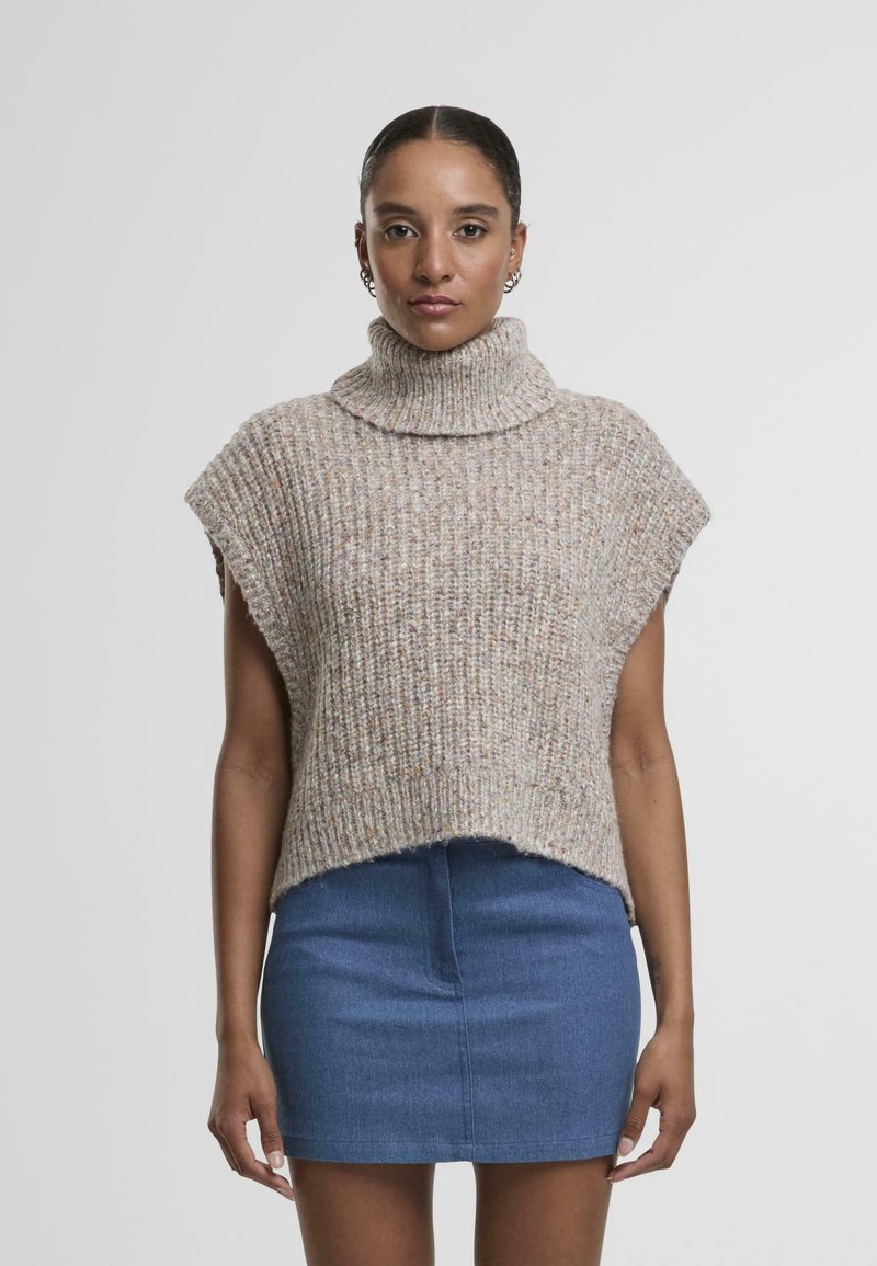 Poncho en tricot côtelé beige, col roulé, manches courtes, porté sur une mini-jupe en denim bleu. L'ourlet est légèrement irrégulier.