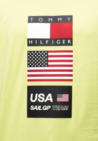 Λογότυπο Tommy Hilfiger πάνω από τη σημαία των Ηνωμένων Πολιτειών και το κείμενο "Ομάδα USA SailGP" με μια μικρή σημαία σε κίτρινο φόντο.