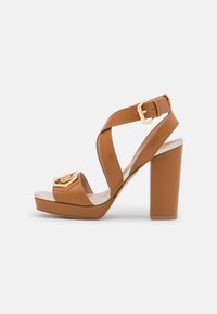 Ruskea keinonahkainen korkokengäsandaali, jossa on ristikkäiset hihnat, neliönmuotoinen kärki, metallinen koriste edessä ja paksu korko lisävakauden takaamiseksi.