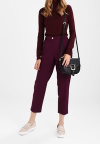 Burgundy turtleneck trøje, højtaljede lilla bukser, beige slip-on sko med frynser og en sort crossbody taske med en guldbuckle.