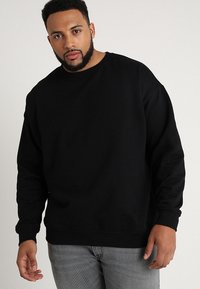 Sudadera negra con cuello redondo, mangas largas y un corte relajado. Tejido suave y puños acanalados, llevada sobre jeans grises.