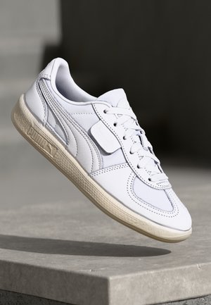 Biały niski sneaker z teksturowaną podeszwą i sznurówkami, umieszczony w powietrzu nad szarą kamienną powierzchnią na ciemnym tle.