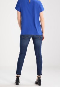 Modrá blůza s krátkým rukávem a detaily klíčové dírky na zádech, spárovaná s tmavými denímovými džíny. Obuv má lesklý černý povrch a mašlový dekor.