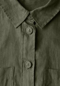 Camicia di lino verde oliva con colletto, due bottoni e taschino frontale in tessuto testurizzato con dettagli cuciti.