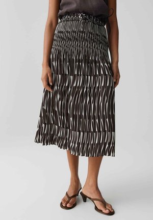 Frau trägt einen schwarz-weißen plissierten Midirock mit abstrakten Mustern und braune Sandalen mit Absatz, steht vor einem einfarbigen Hintergrund.