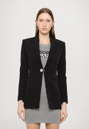Blazer negro a medida con solapa estructurada y cierre de un solo botón plateado, usado sobre un vestido de rayas gris y negro. Textura de tela suave.