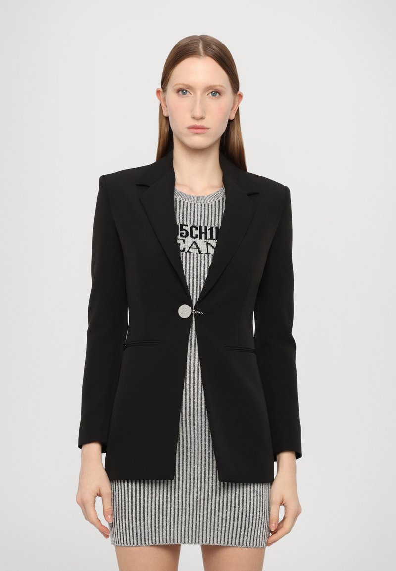 Blazer nero su misura con revers strutturato e chiusura con un unico bottone argentato, indossato sopra un vestito a righe grigie e nere. Tessuto dalla trama liscia.