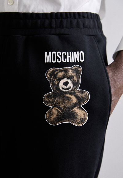 Pantaloni negri de trening cu un grafic texturat de urs pe coapsă, având brandingul alb "Moschino" deasupra imaginii.