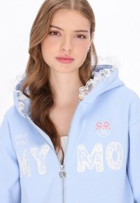 Sudadera con capucha azul claro con cremallera, capucha con borde de encaje blanco, palabras bordadas "MY MO" y texto "Llena de energía". Textura de tela suave.