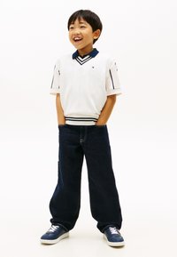 Χαμογελαστός νεαρός αγόρι που φοράει ένα λευκό αμάνικο πουλόβερ πάνω από ένα μπλε σκούρο πουκάμισο, με τα χέρια στις τσέπες των σκούρων μπλε τζιν και μπλε σκούρα αθλητικά παπούτσια.