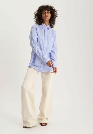 Femme aux cheveux bouclés portant une chemise bleue rayée oversize et un pantalon crème à jambes larges, debout pieds nus sur un fond uni.