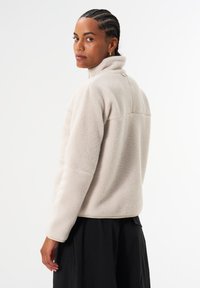 Fleecepullover in Hellbeige mit hohem Kragen, einer Textur, die weichem Lammfell ähnelt, und kontrastierendem Besatz entlang des Saums und der Bündchen.