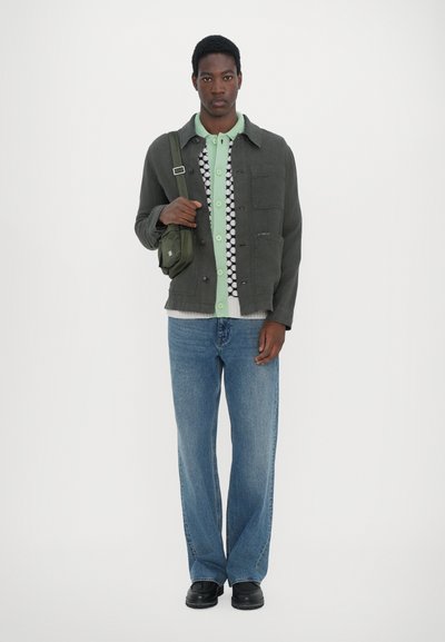 Veste verte sur une chemise à motifs avec boutons, associée à un jean large bleu clair et des chaussures noires. Un sac bandoulière vert pendre de l'épaule.
