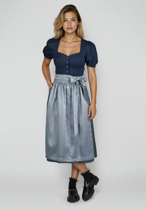 Frau trägt eine marineblaue Bluse mit Puffärmeln und einen silbernen Satinschürzenrock, dazu schwarze Socken und Plateauloafer, stehend vor einem einfarbigen Hintergrund.