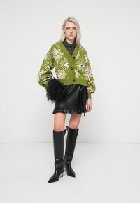 Cardigã verde de malha com padrões florais, saia preta de imitação de couro com babados e botas pretas de bico alto até ao joelho.