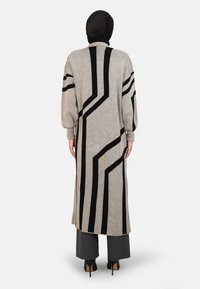 Long, gray cardigan with black geometric stripes, knit texture, high collar, and dropped sleeves, worn over gray pants and heels.
