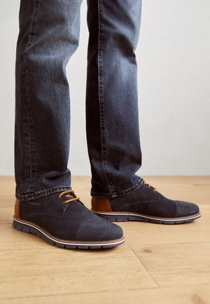Zapatos de ante azul marino para hombre con acentos en color marrón claro, que cuentan con una puntera de tela texturizada y cordones, complementados por una suela blanca en contraste y una suela de goma.
