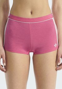 Femme portant un short Adidas rose avec des rayures blanches sur les côtés, debout avec les mains détendues sur les côtés.