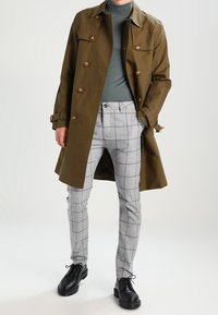 Płaszcz trencz w oliwkowej zieleni z dużymi guzikami, szare spodnie w kratę oraz czarne buty. Strój uzupełnia golf pod płaszczem.