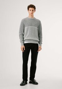 Grau gestrickter Pullover mit strukturiertem Muster, Rundhalsausschnitt und gerippten Bündchen, kombiniert mit schwarzen Hosen und schwarzen Schuhen.