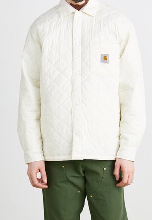 Homme portant une veste matelassée blanche à boutons avec une poche poitrine ornée d'un logo Carhartt et un pantalon vert olive.