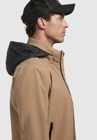 Chaqueta beige para hombre con capucha negra, con cierre frontal con cremallera, bolsillos laterales y textura suave. Ajuste clásico con elementos de diseño modernos.