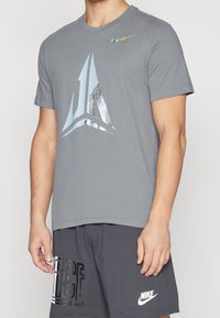 La camiseta gris de manga corta presenta un logotipo geométrico reflectante y un pequeño logotipo en el hombro. Está hecha de una suave mezcla de algodón.