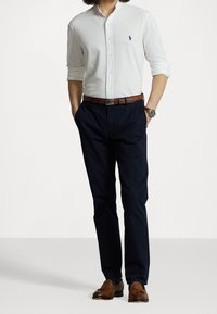 Camisa blanca de botones con mangas arremangadas y un logo azul, combinada con pantalones azul marino y mocasines de cuero marrón. Cinturón marrón acentúa el conjunto.