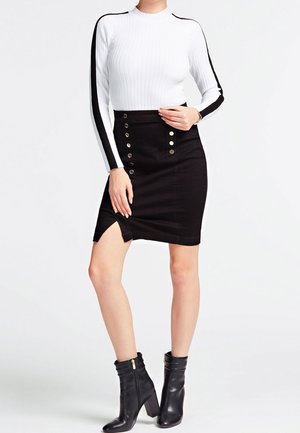 Femme portant un pull blanc côtelé avec des rayures noires et une jupe taille haute noire avec des boutons et une fente latérale, associée à des bottines noires à talons.