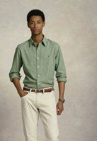 Zaļš krekls ar pogām un apkakli, kas valkāts virs krēmkrāsas biksēm. Satur divas priekšējās kabatas un mazu logotipu uz krūts.