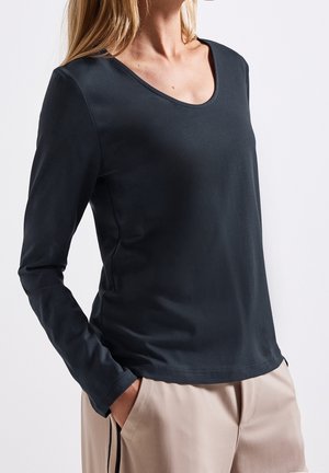 Vrouw draagt een zwart strakvallend shirt met lange mouwen en een ronde hals, gecombineerd met beige broek, staand met handen in de zakken tegen een witte achtergrond.