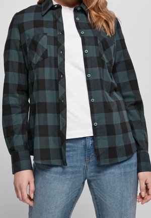 Femme portant une chemise à carreaux noire et vert foncé déboutonnée sur un t-shirt blanc et un jean bleu, sur un fond uni.