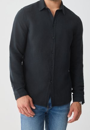 Man draagt een zwart, lang mouwen, overhemd met knopen en blauwe spijkerbroek, staand tegen een effen lichte achtergrond.