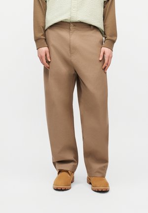 Person, die locker sitzende braune Hose, beige Stiefel und ein hellgrünes, gemustertes Oberteil mit braunen Ärmeln trägt, vor einem weißen Hintergrund steht.
