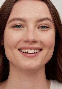 Nærbilde av en smilende person med lys hud, lys brunt hår, blå øyne og rette tenner. Har naturlig sminke og nøytral bakgrunn.