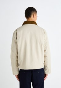 Chaqueta de algodón beige con un collar de pana marrón, corte recto y botones decorativos en la parte posterior, combinada con pantalones oscuros.