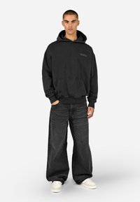 Melna kapuce ar priekšējo kabatu un izšūtu logo, kombinēta ar brīvā stila melnajām džinsām un baltām kedām. Ikdienišķs ielu apģērbu dizains.