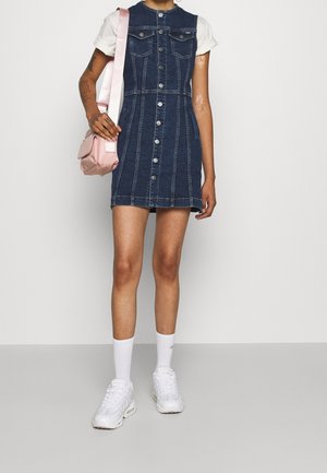 Vestido de mezclilla en azul oscuro con cierre de botones, dos bolsillos en el pecho y una silueta ajustada. Combinado con un bolso cruzado rosa y zapatillas blancas.