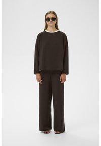 Conjunto de ropa de estar en color marrón que incluye una parte superior de manga larga de corte holgado con un cuello blanco y pantalones de pernera ancha, fabricado en una suave tela. Se llevan gafas de sol.