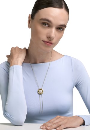 Jonge vrouw in een lichtblauwe top met lange mouwen die een gouden hanger met een centrale parel draagt, met de ene hand op haar schouder en de andere op een wit oppervlak rustend.