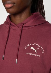 Bordeaux hettegenser med snorer, med et kremfarget logo som leser "PUMA ATHLETICS" og "etablert 1948" med en hoppende puma-design.