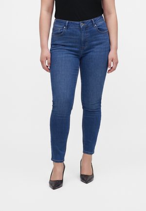 Femme portant un jean skinny bleu et des talons noirs à bouts pointus, debout les mains détendues le long du corps sur un fond uni.