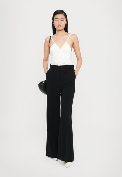 Haut blanc ajusté avec des détails froncés, associé à un pantalon noir à taille haute et à jambes larges, et des talons aigus blancs ; tient un sac à bandoulière noir.