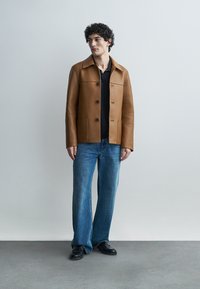 Giovane uomo con capelli ricci che indossa una giacca di pelle marrone, una maglietta nera, jeans blu e scarpe nere, in piedi contro uno sfondo semplice di colore grigio chiaro.