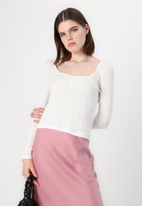 Haut blanc à manches longues avec des rayures texturées et décolleté carré, associé à une jupe en satin rose. La silhouette tient un chouchou fleuri noir.