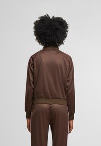 Ruda zip-up striukė su tekstūruotu raštu, raukšlėtais rankogaliais ir apdaila. Yra apykaklė ir subtilūs šoniniai akcentai. Lygus, lengvas audinys.