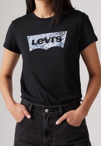 Kvinna i en svart Levi's t-shirt med en blåmönstrad logotyp, instoppad i svart högmidjad jeans, med händerna i fickorna.